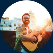 Artista tocando guitarra acústica al aire libre durante el atardecer, disfrutando de la música con una sonrisa en un entorno urbano. Inspirador fondo de edificios y puesta de sol.